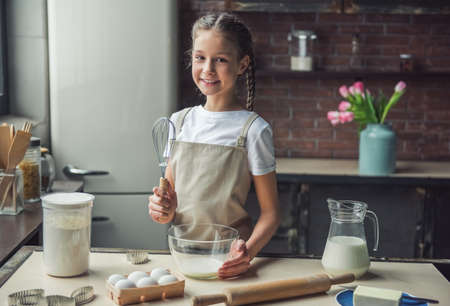 Cute little girl in apron is holding a whisk, looking at camera and smiling while preparing dough in kitchen at homeの写真素材