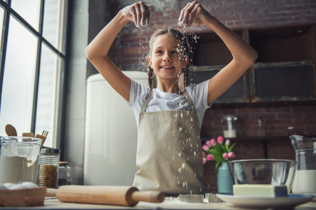 Cute little girl in apron is playing with flour and smiling while cooking in kitchen at home