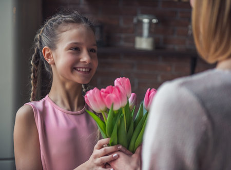 Little girl is giving flowers to her mom and smiling while standing in kitchen at homeの写真素材