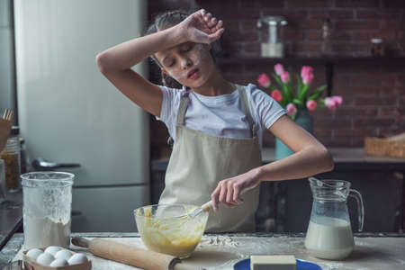 Cute tired little girl in apron is using a whisk and wiping her forehead while preparing dough, in kitchen at homeの写真素材