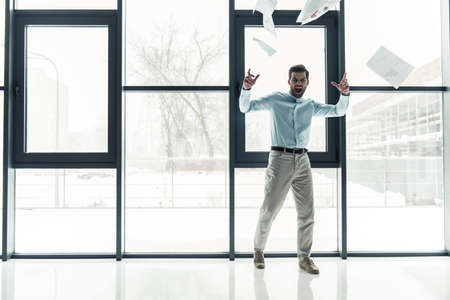 Handsome emotional businessman is throwing papers and screaming while standing near the window in the office buildingの写真素材