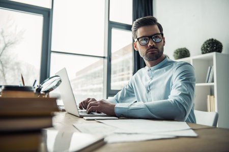 Handsome pensive businessman in eyeglasses is looking away and thinking while working with the laptop in the officeの写真素材