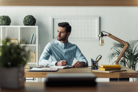 Handsome pensive businessman is making notes and looking away while working in the office