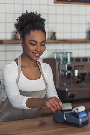 Beautiful Afro American barista in apron is using a payment terminal and smiling while standing at...