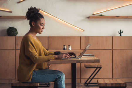 Side view of beautiful Afro American girl in casual clothes smiling while working with a laptop in cafeの写真素材