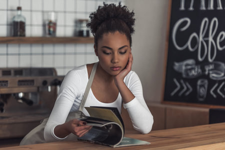 Beautiful Afro American bored barista in apron is reading a magazine while leaning on the bar...