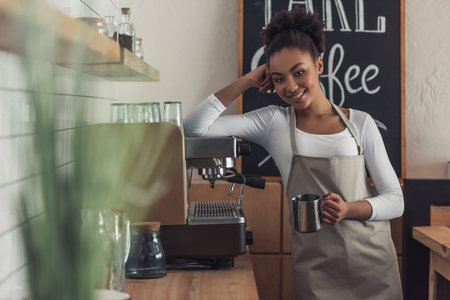 Beautiful Afro American barista in apron is holding a cup, looking at camera and smiling while leaning on the coffee machineの写真素材