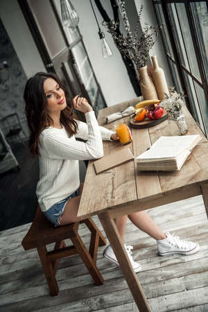 Beautiful young girl in casual clothes is writing in the notepad, holding a glass of juice and smiling while sitting at the wooden table at homeの写真素材