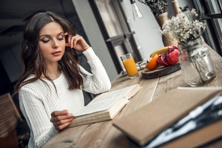 Beautiful young girl in casual clothes is reading a book while sitting at the wooden table at home
