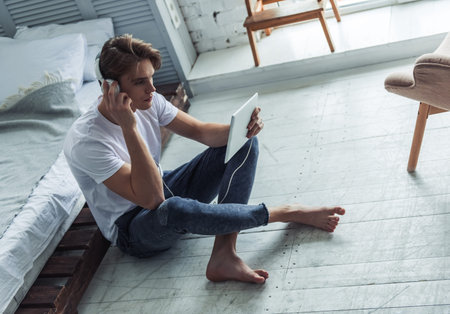 Handsome young man in casual clothes is using a digital tablet and headphones while sitting on the...
