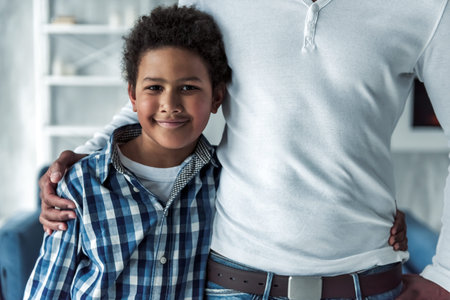 Happy Afro American boy in casual clothes is looking at camera and smiling while hugging his dad