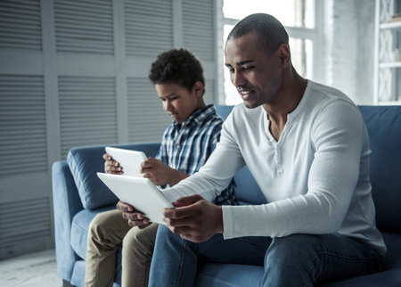 Happy Afro American father and son in casual clothes are using digital tablets and smiling while...