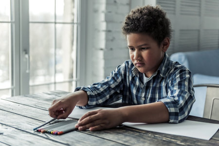 Sad Afro American boy in casual clothes is collecting colored pencils while sitting at the table at homeの写真素材