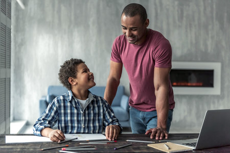Handsome Afro American man is watching his son drawing, both are smiling, at homeの写真素材