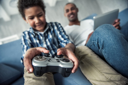 Afro American father and son in casual clothes using gadgets, sitting on couch at home, boy is playing video game console in focusの写真素材