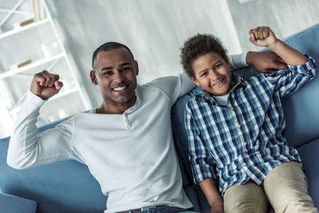 Happy Afro American father and son in casual clothes are raising their fists, looking at camera and smiling while sitting on couch at homeの写真素材
