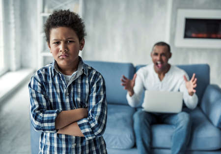 Afro American boy is holding arms crossed and looking offendedly at camera while his dad is screaming, sitting on couch in the backgroundの写真素材