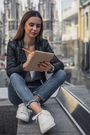 Beautiful young girl in casual clothes is making notes and smiling while resting in city centerの写真素材