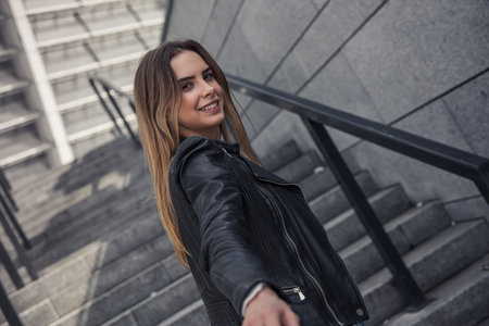 Beautiful young girl in casual clothes is stretching her hand, looking at camera and smiling while...
