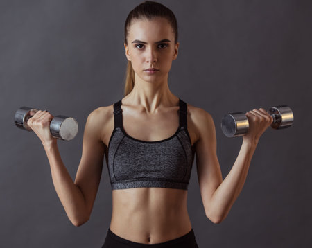 Beautiful girl in sportswear is doing exercises using dumbbell and looking at camera, on gray...