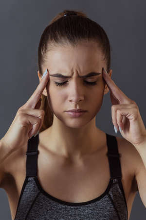 Beautiful girl in sportswear is touching her temples feeling pain, on gray backgroundの写真素材