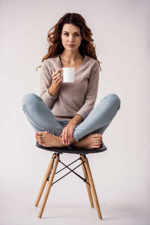 Portrait of a young beautiful girl holding a cup in her hand that drinks tea or coffee, sitting on a chair and smiling at the camera on a white backgroundの写真素材