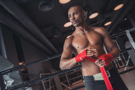 Afro American boxer is wrapping hands with red bandage and looking away while preparing to train in gymの写真素材