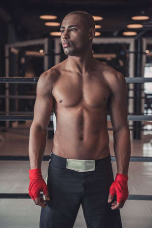 Afro American boxer, his hands wrapped in red bandage, is looking away while standing in pose in gymの写真素材