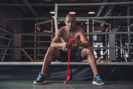 Afro American boxer is wrapping hands with red bandage and looking at camera while sitting on the boxing ringの写真素材