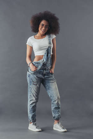 Afro American woman in jean overalls and eyeglasses is posing at camera and smiling, on a grey background, full-lengthの写真素材