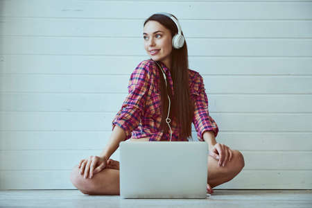 Portrait of a young beautiful girl with long hair uses a laptop and listens to music in headphones while sitting on the floorの写真素材