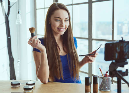 Young beautiful woman blogger using cosmetics while shooting at camera at homeの写真素材
