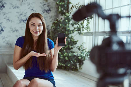 Young woman blogger makes presentation of her phone in front of camera at homeの写真素材