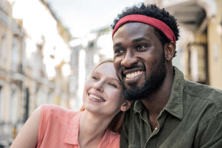 Young cheerful couple where a black man and a blonde woman are smiling embracing while sitting at a table in a cafe while walking through the city streetsの写真素材