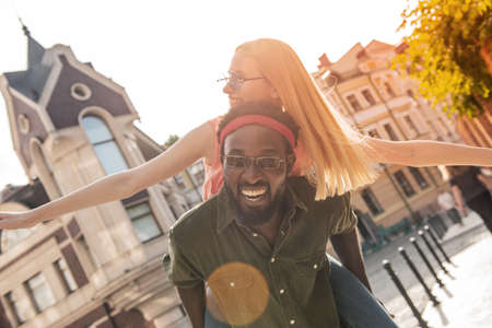Young cheerful couple where a black man and a blonde woman are smiling. Girl sitting on the back of a man with his arms outstretched while walking around the cityの写真素材
