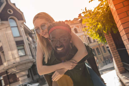 Young cheerful couple where a black man and a blonde woman are smiling. Girl sitting on the back of a man hugs his neck while walking around the cityの写真素材