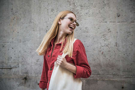 Young cheerful blonde girl in sunglasses and red jacket posing near a gray wallの写真素材