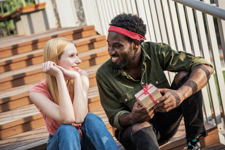 A young black man gives a gift to his blonde girlfriend sitting on the stairs while walking