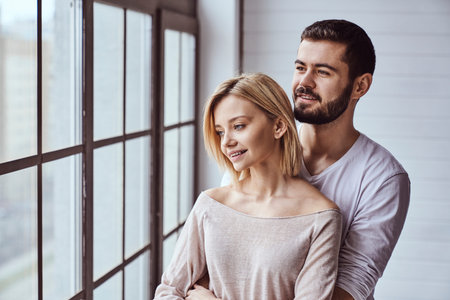 Portrait of young cheerful loving couple smiling looking at camera and hugging each other looking out the window at homeの写真素材