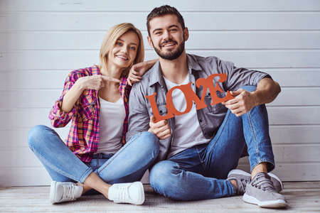 Young happy loving couple sitting on floor and holding sign "Love", smiling at cameraの写真素材