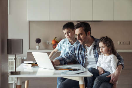 Father at work remotely with a laptop and documents sitting at a table at home in the kitchen entertains his little daughter and sonの写真素材