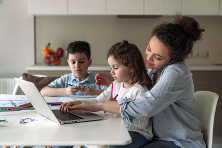 Mom at home with computer and documents sitting at the table in the kitchen and her little daughter and son drawing with colored pencils and markersの写真素材