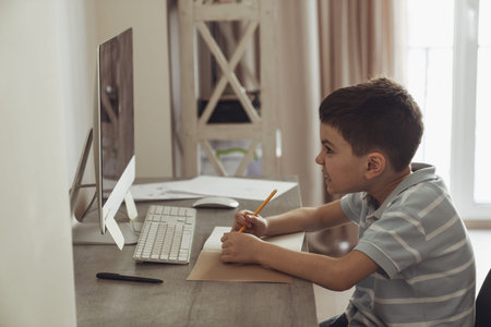 Little boy sitting in his room at a desk next to a computer and doing homework preparing for school