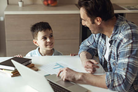 Father at work remotely with laptop sitting at table at home in the kitchen and his little son wanting attentionの写真素材