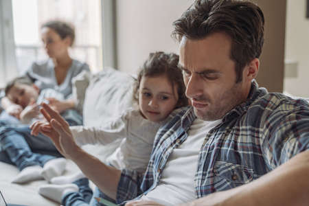 A young successful freelancer man works at home sitting on the couch against the backdrop of his wife and young son and daughter, who are currently watching TV and having fun, the daughter wants attention, what prevents him from workingの写真素材