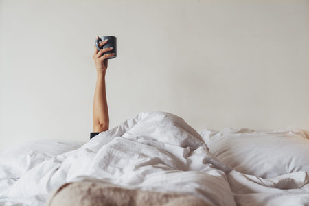 The hand of a young woman who can't wake up holding a mug of coffee or tea from under a blanket in...
