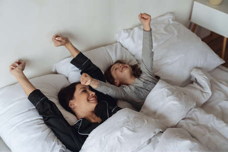 Mom and daughter waking up after sleeping in their bed together and stretching their arms up in the bedroom at home.の写真素材