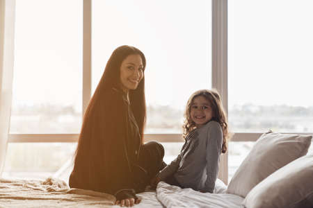Mom and daughter woke up in the morning sitting on the bed in front of a large window.の写真素材