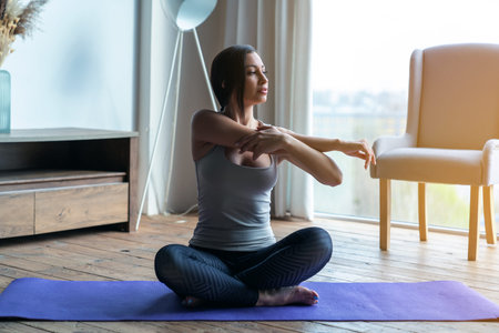Young beautiful fitness woman at home sitting in lotus position on mat during yoga class in living...