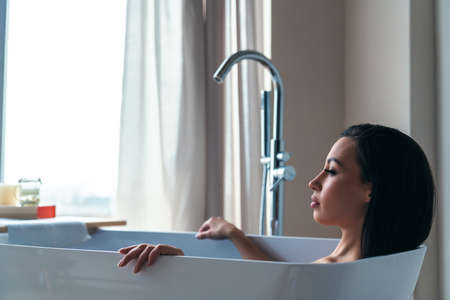 Young beautiful woman relaxes taking a bath in the bathroom at home on the background of a large window.の写真素材
